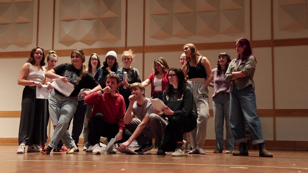 A still from "Premium Orange." A group of mostly-white college students pose onstage during a rehearsal.