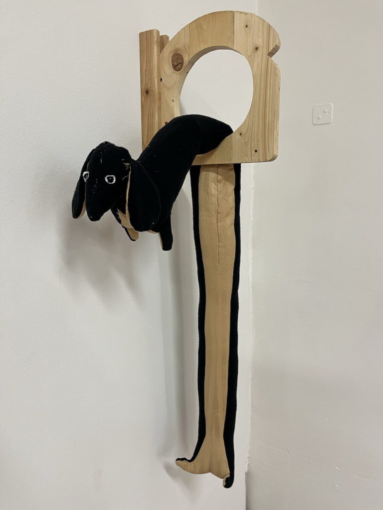 A wooden piece of toast with a hole in it; a long fabric dog is draped through the hole